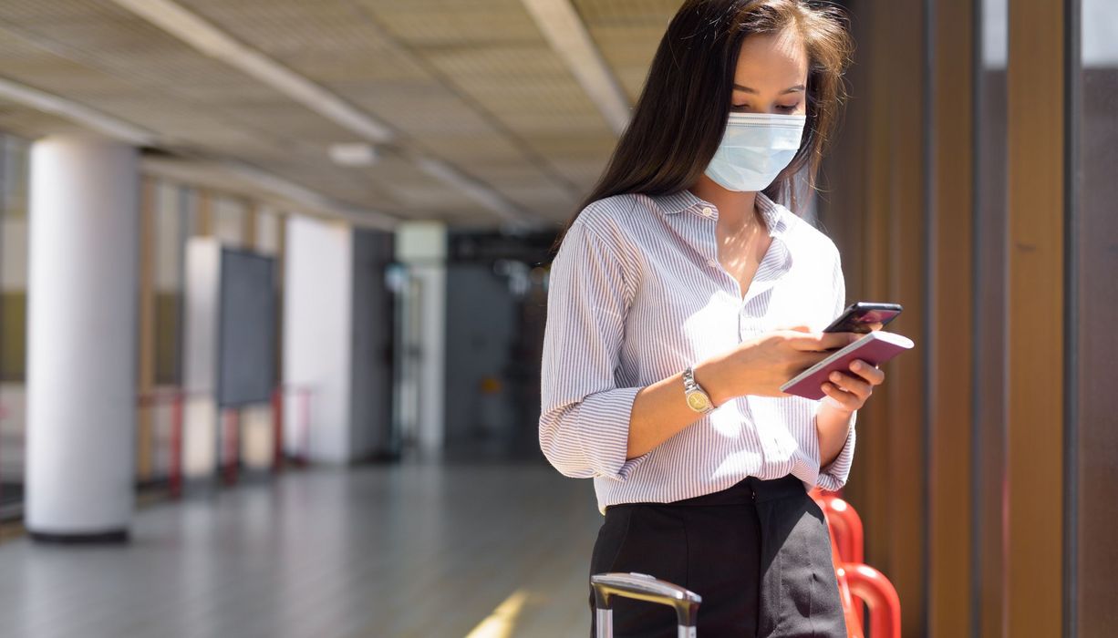 Junge Frau am Flughafen mit Maske, Koffer und Smartphone.