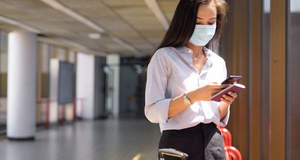 Junge Frau am Flughafen mit Maske, Koffer und Smartphone.