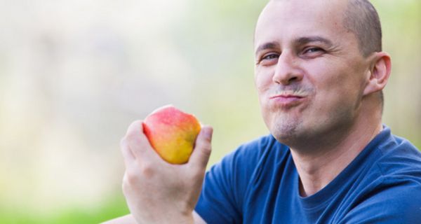 Mann mit kurzen Haaren und blauem T-Shirt kaut einen Bissen Apfel und hält den Apfel in der Hand