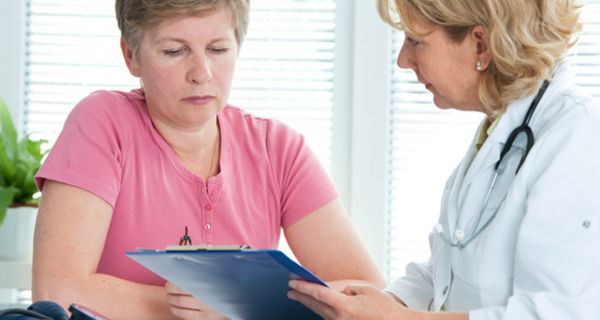 Patientin in mittleren Jahren mit Ärztin, die ein Clipboard in der Hand hält und ihr Fragen stellt.