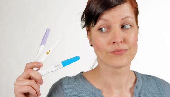 Jüngere Frau mit zusammengebundenen, braunen Haaren, grauer Pulli, schaut skeptisch auf 3 Schwangerschaftstests in ihrer rechten Hand
