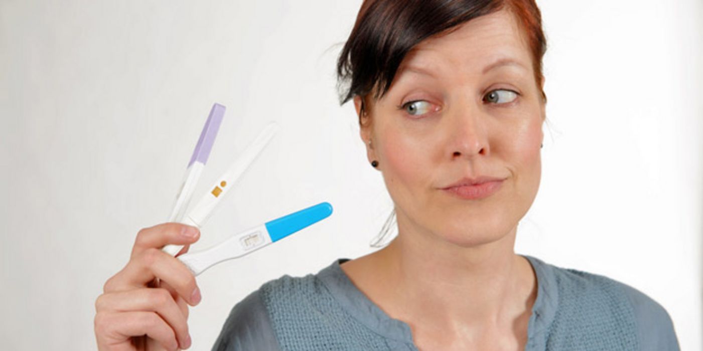 Jüngere Frau mit zusammengebundenen, braunen Haaren, grauer Pulli, schaut skeptisch auf 3 Schwangerschaftstests in ihrer rechten Hand