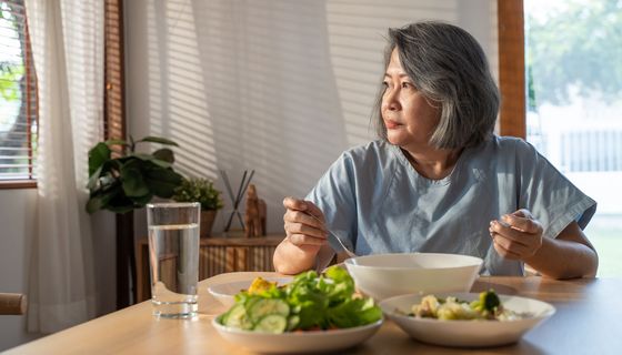 Ältere Frau, schaut traurig aus dem Fenster und isst allein.
