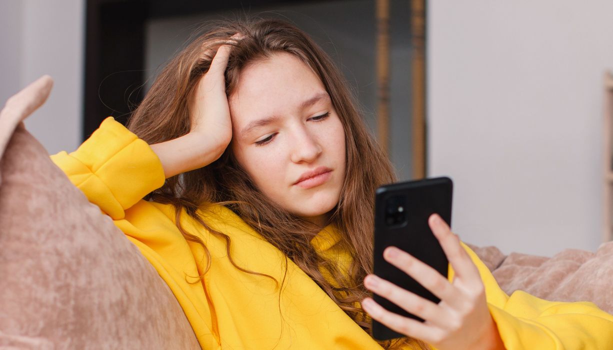 Jugendliche sitzt auf dem Sofa und ist am Smartphone. Sie blickt mäßig glücklich.