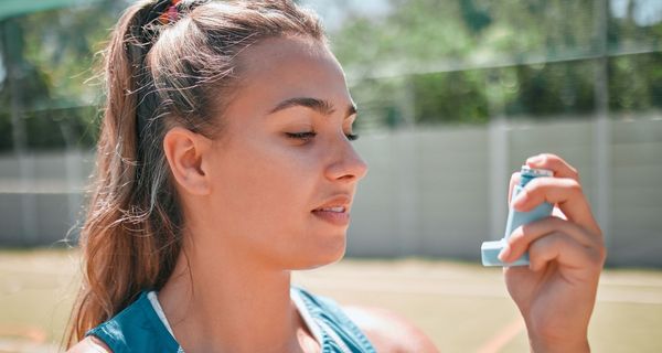 Junge Frau auf einem Sportplatz in Sportkleidung blickt auf den Asthma-Inhalator in ihrer Hand.