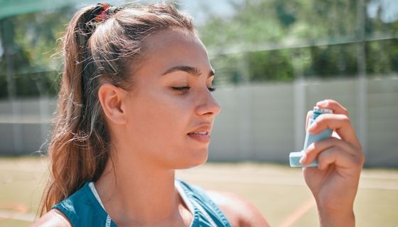 Junge Frau auf einem Sportplatz in Sportkleidung blickt auf den Asthma-Inhalator in ihrer Hand.
