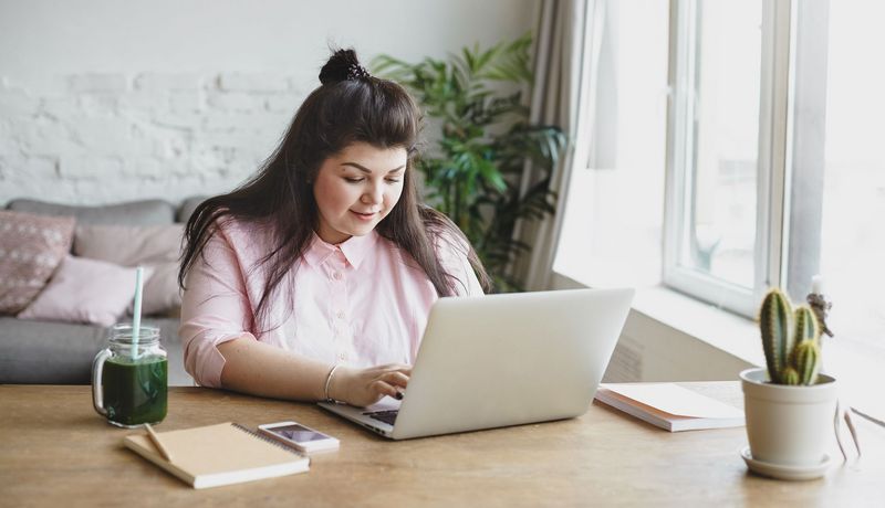 Eine übergewichteige junge Frau sitzt vor einem Laptop und tippt.