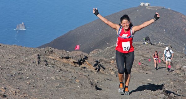 Ultra-Marathon-Läuferin auf Berggipfel, im Hintergrund das Meer, reißt freudig die Arme hoch