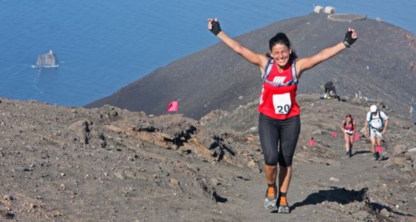 Ultra-Marathon-Läuferin auf Berggipfel, im Hintergrund das Meer, reißt freudig die Arme hoch