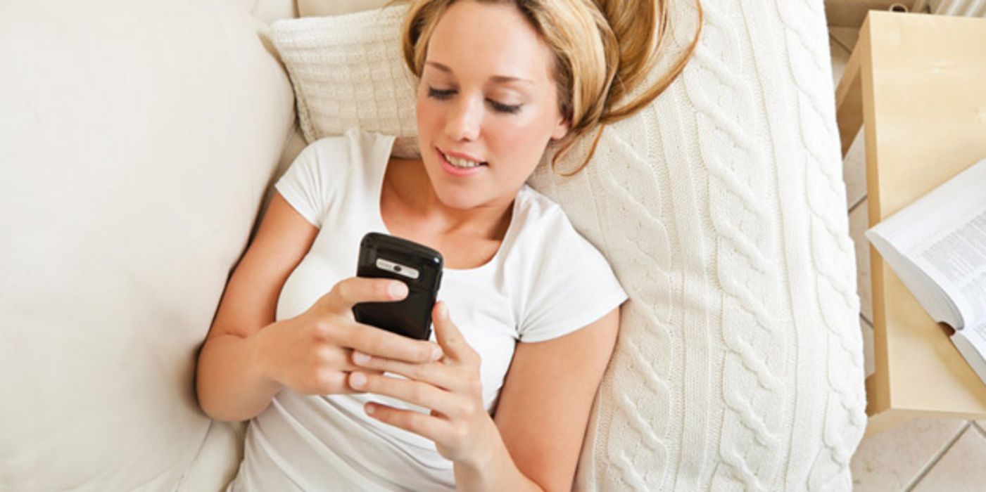 Junge blonde Frau liegt im Bett auf dem Rücken und schaut lächelnd in ihr Smartphone