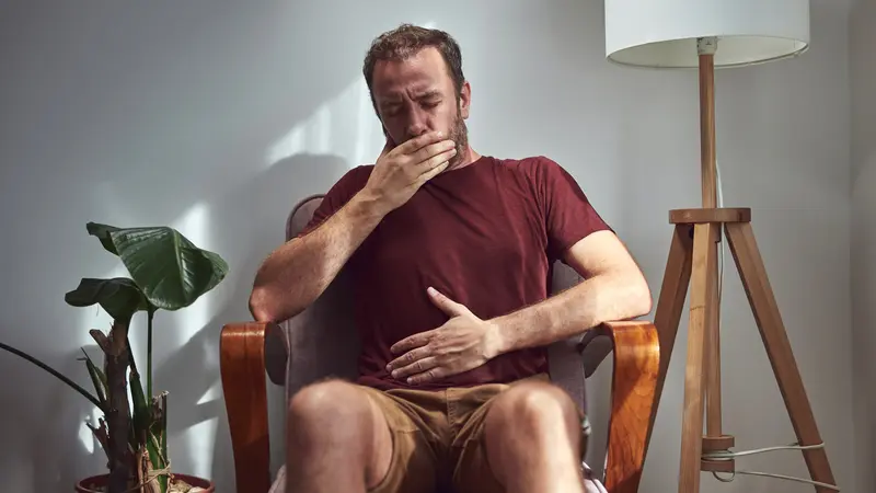 Erwachsener Mann sitzt auf einem Sessel zuhause, hält sich den Bauch und die Hand vor den Mund.
