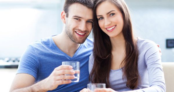 Junges sympathisches Paar lacht in die Kamera, jeweils ein Wasserglas in der Hand