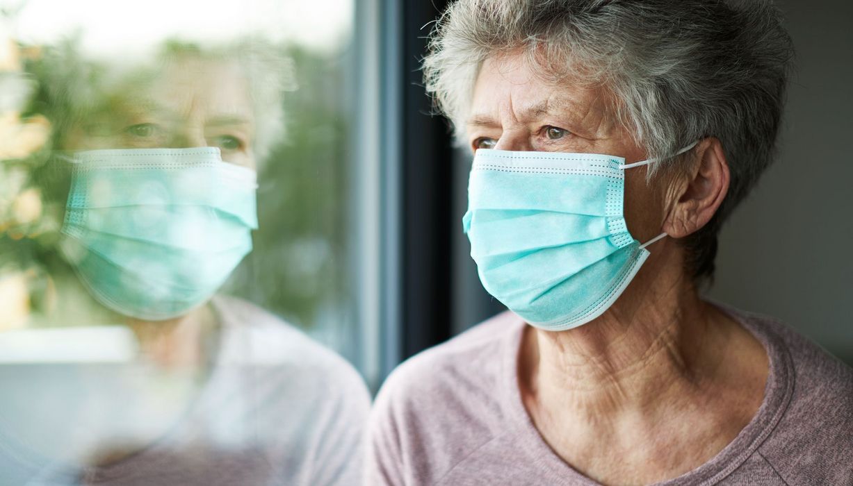 Seniorin mit Maske, schaut aus dem Fenster.