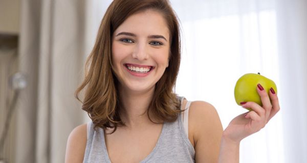 Dunkelhaarige strahlend lachende junge Frau (frontal zur Kamera, graues, ärmelloses T-Shirt), in der linken Hand einen grünen Apfel haltend