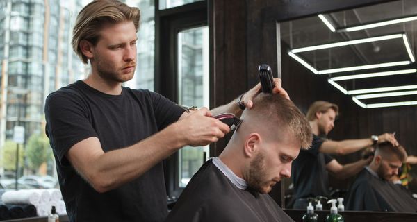 Ein Mann steht in einem Salon und rasiert einem anderen Mann den Kopf. Dieser hat den Kopf leicht nach vorne gebeugt.