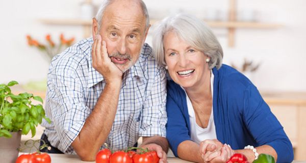 Vergnügtes Seniorenpaar mit Tomaten und Paprika auf Tisch lachen in die Kamera, links Zinktopf mit Basilikum