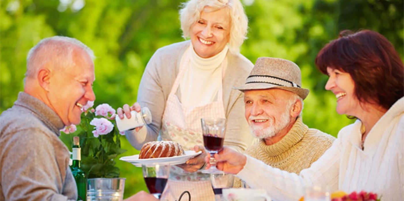 Sommer: Geburtstagsfeier draußen: 2 fröhliche Seniorenpaare feiern mit Geburtstagskuchen und Rotwein, stoßen gerade an mit Geburtstagskind