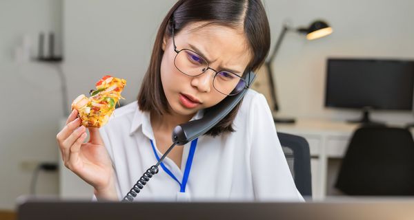 Junge Frau telefoniert, liest gleichzeitig etwas auf einem Bildschirm und hält einen Pizza-Snack in der Hand. Im Hintergrund: Ein dunkler Arbeitsplatz.
