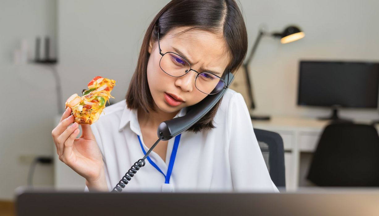 Junge Frau telefoniert, liest gleichzeitig etwas auf einem Bildschirm und hält einen Pizza-Snack in der Hand. Im Hintergrund: Ein dunkler Arbeitsplatz.