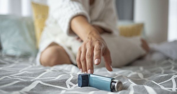 Junge Frau, sitzt im Bett und greift nach Asthmaspray.