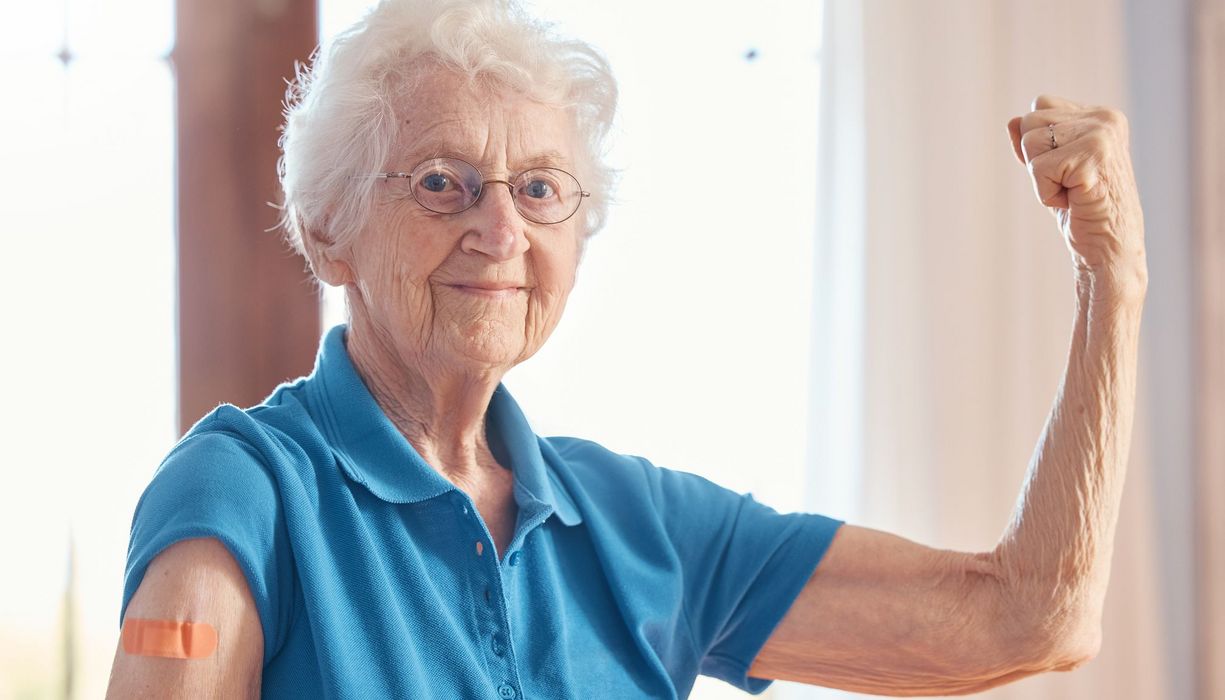 Ältere Dame mit weißem Haar und Brille präsentiert ihren Oberarm mit Pflaster. Gleichzeitig spannt sie ihren anderen Arm an und blickt stolz in die Kamera.