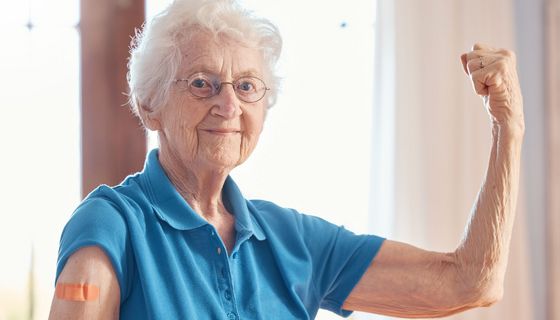 Ältere Dame mit weißem Haar und Brille präsentiert ihren Oberarm mit Pflaster. Gleichzeitig spannt sie ihren anderen Arm an und blickt stolz in die Kamera.