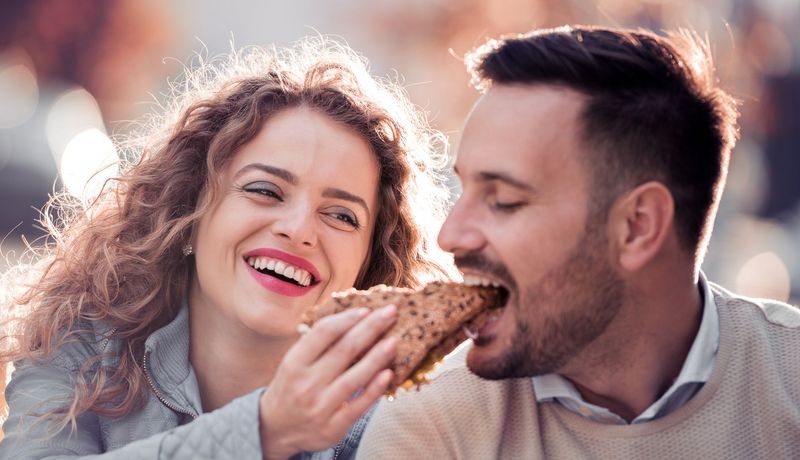 Junge Frau lässt einen Mann von ihrem Vollkorn-Brötchen abbeißen.