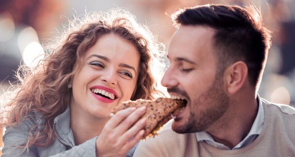Junge Frau lässt einen Mann von ihrem Vollkorn-Brötchen abbeißen.