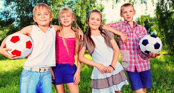 Zwei Jungen und zwei Mädchen stehen in sommerlichen Outfits im Freien, die Jungen tragen je einen Fußball unter dem Arm