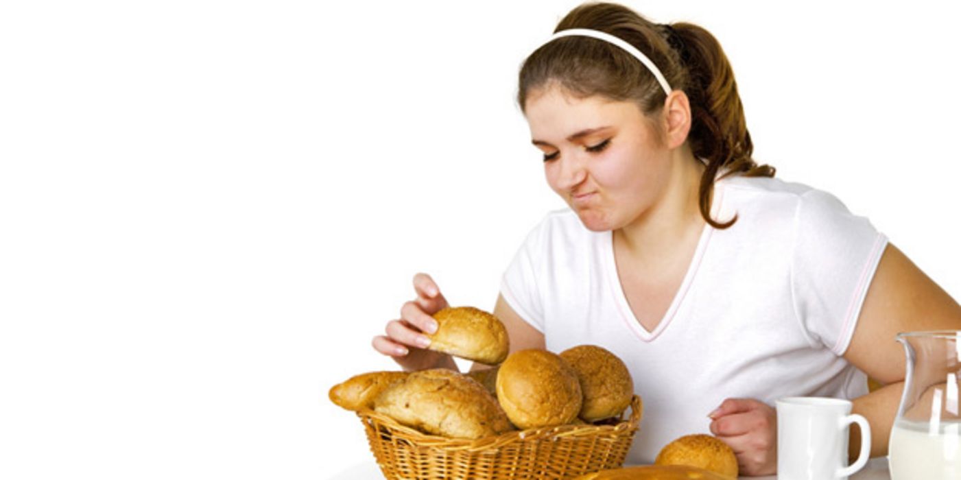 Leicht übergewichtige junge Frau greift zögerlich nach einem Brötchen aus einem Brotkorb