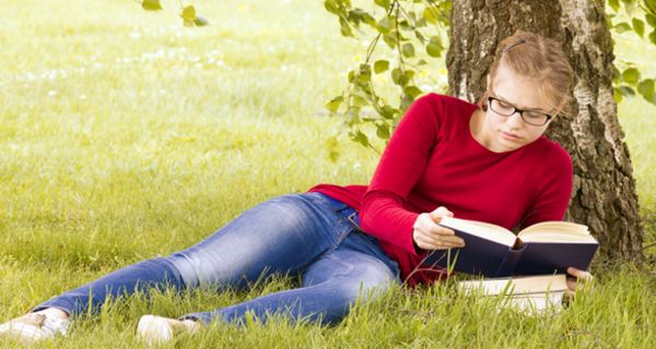 Junge Frau liegt unter einer Birke im Gras und liest ein Buch.