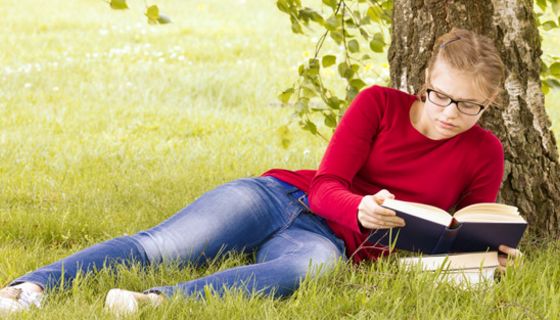 Junge Frau liegt unter einer Birke im Gras und liest ein Buch.