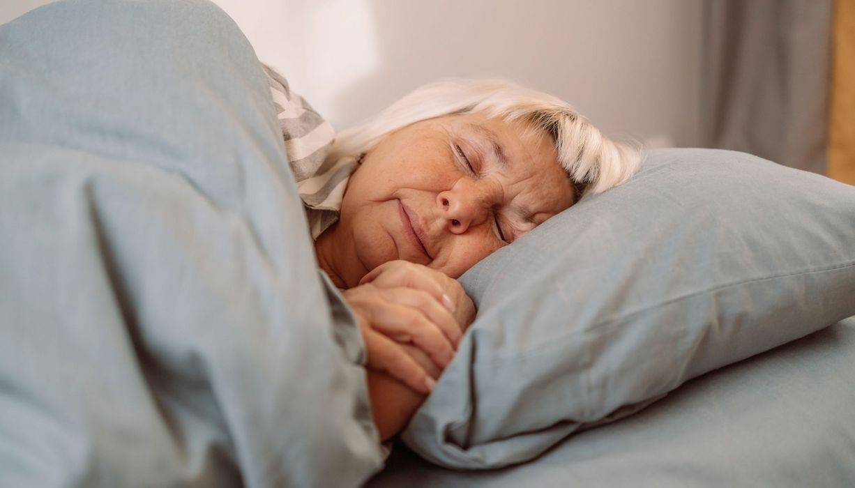 Eine ältere Dame mit blondiertem Haar schläft in grauer Bettwäsche.