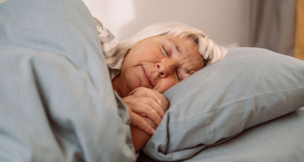 Eine ältere Dame mit blondiertem Haar schläft in grauer Bettwäsche.