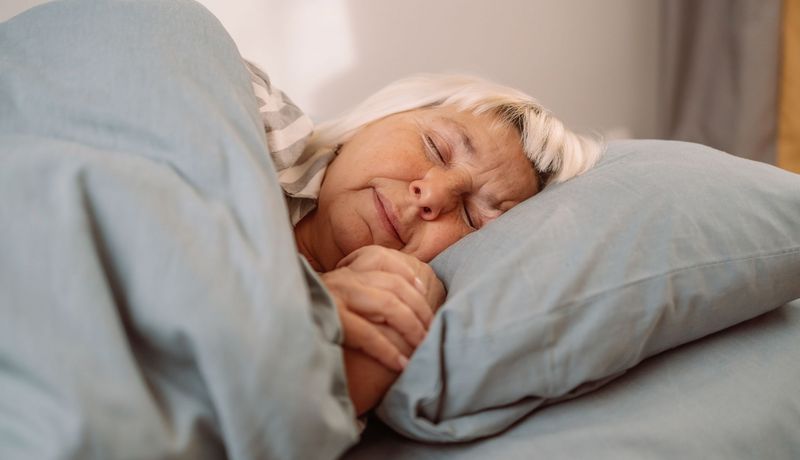Eine ältere Dame mit blondiertem Haar schläft in grauer Bettwäsche.