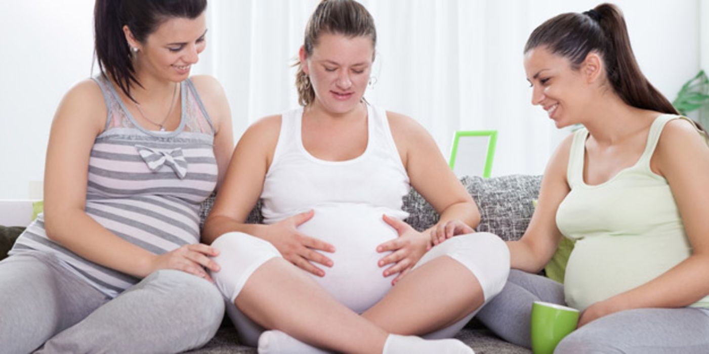 Drei Hochschwangere sitzen in Leggins und ärmellosen Shirts auf einem Sofa, die mittlere hat die Hände auf dem Bauch, die beiden rechts und links schauen dorthin.