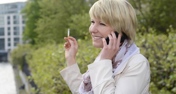 Frau in den 40ern, Profilbild, blond, Zigarette, Handy, vor Busch, Hintergrund Häuser, heller Trenchcoat