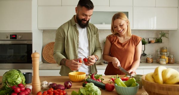 Ein Paar schneidet zusammen Gemüse. Auf der Arbeitsfläche liegen Paprika, Tomaten, Salat, Zwiebeln und vieles mehr.