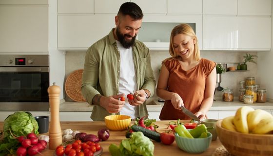 Ein Paar schneidet zusammen Gemüse. Auf der Arbeitsfläche liegen Paprika, Tomaten, Salat, Zwiebeln und vieles mehr.