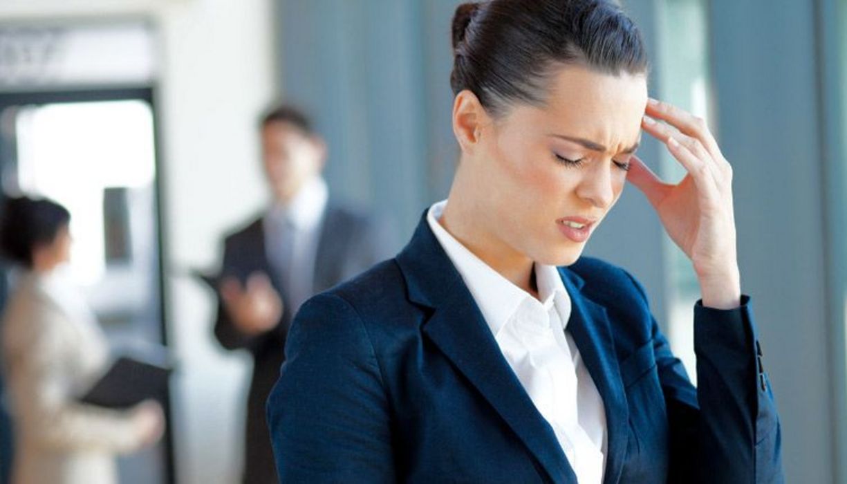 Büroszene: Jüngere Frau im Vordergrund, Bild Oberkörper, Businesskleidung (dunkelblaues Jacket, weiße Bluse, zurückgekämmte, dunkle Haare) greift sich an die schmerzende Stirn, im Hintergrund unscharf Frau und Mann, Büro