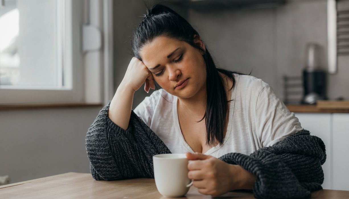 Frau, sitzt müde mit einer Tasse Kaffee am Tisch.