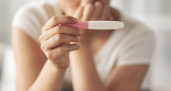 Im Vordergrund: Hand hält einen Schwangerschaftstest (Ergebnis nicht sichtbar), im Hintergrund (unscharf): Eine Frau, die besorgt wirkt, die Hand am Mund. 