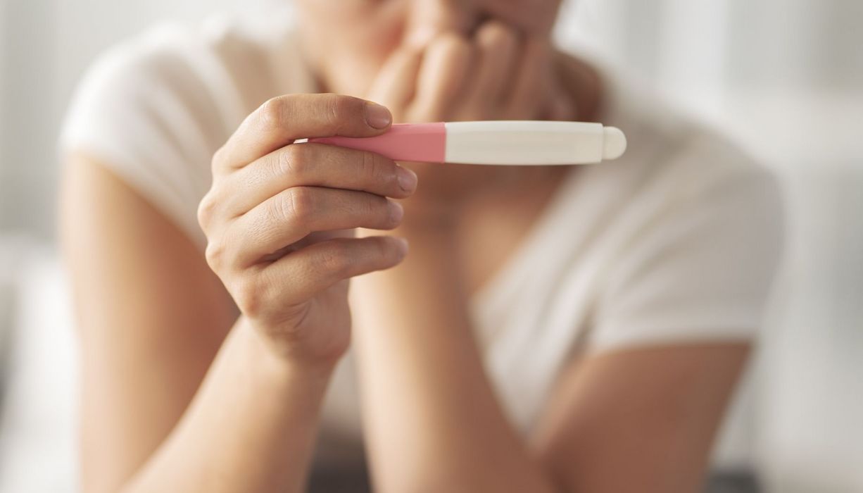 Im Vordergrund: Hand hält einen Schwangerschaftstest (Ergebnis nicht sichtbar), im Hintergrund (unscharf): Eine Frau, die besorgt wirkt, die Hand am Mund. 