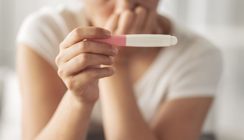 Im Vordergrund: Hand hält einen Schwangerschaftstest (Ergebnis nicht sichtbar), im Hintergrund (unscharf): Eine Frau, die besorgt wirkt, die Hand am Mund.