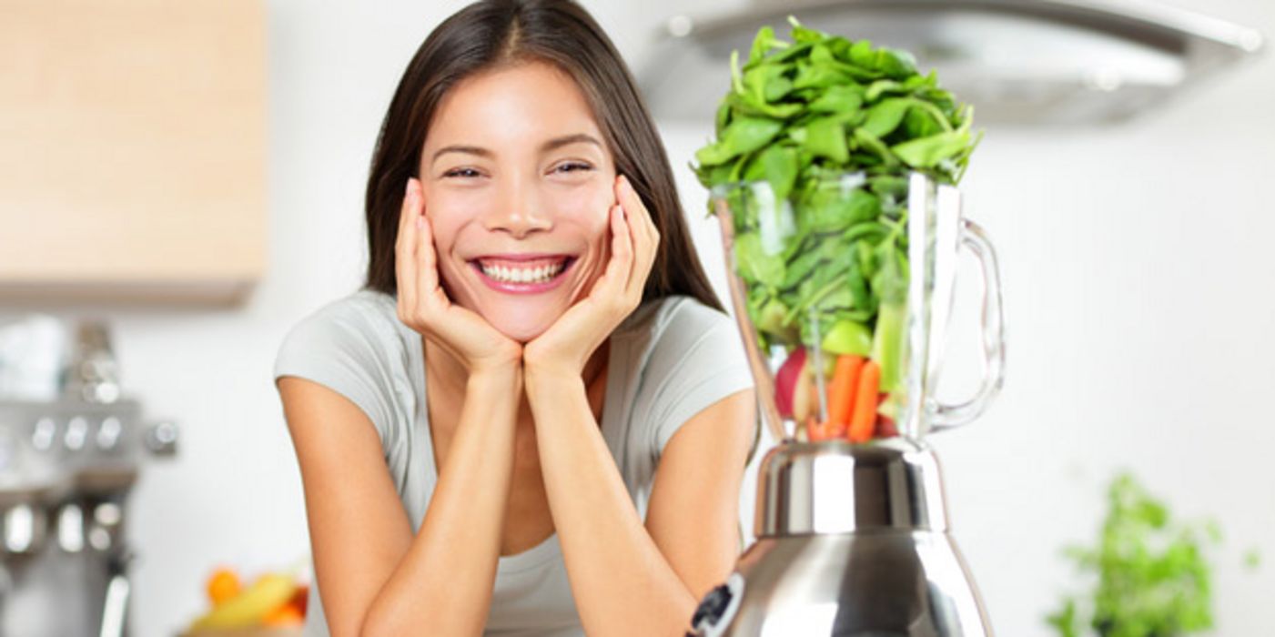 Strahlende junge Frau (unser aponet-Topmodel), Gesicht in die Hände gestützt, an weißer Küchenarbeitsplatte, darauf ein Mixer mit verschiedenen Gemüsesorten