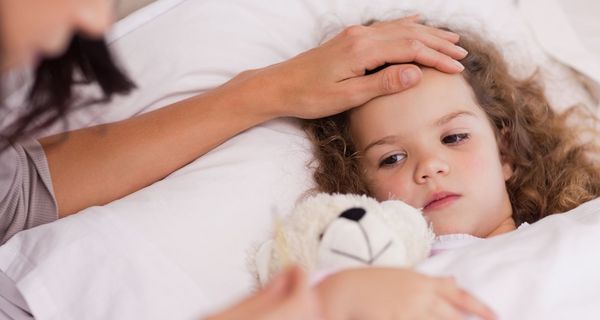 Krankes Kind liegt mit Teddy-Bär im Bett. Jemand legt ihm eine Hand auf die Stirn.