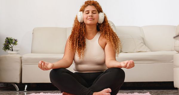 Frau sitzt in Zimmer auf einem Teppich vor einer Couch. Sie trägt Kophörer und hat geschlossene Augen.