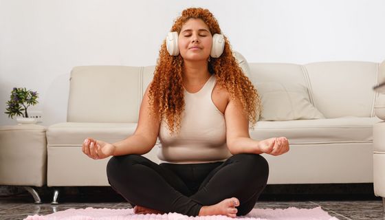 Frau sitzt in Zimmer auf einem Teppich vor einer Couch. Sie trägt Kophörer und hat geschlossene Augen.