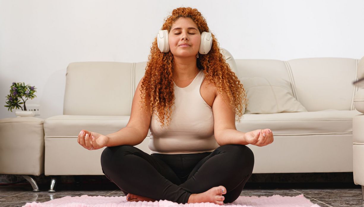 Frau sitzt in Zimmer auf einem Teppich vor einer Couch. Sie trägt Kophörer und hat geschlossene Augen. 