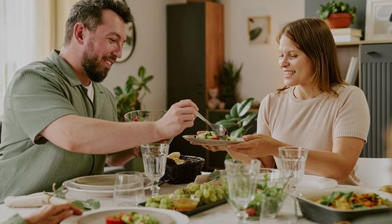 Ein Mann häuft einer Frau Salat auf den Teller, beide wirken glücklich. Auf dem Tisch steht pflanzenbetontes Essen: Salat, Trauben und ein Auflauf.